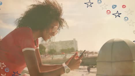 Coloridos-Iconos-De-Estrellas-Contra-Una-Mujer-Afroamericana-Usando-Un-Teléfono-Inteligente-Cerca-De-La-Playa
