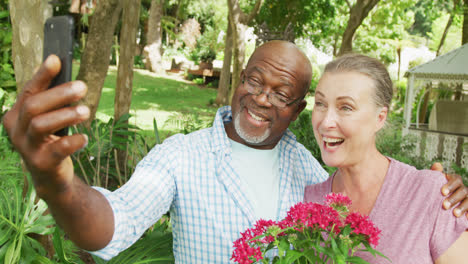 Happy-senior-diverse-couple-wearing-shirts-and-taking-selfie-with-smartphone-in-garden