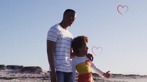Animation-of-hearts-over-african-american-father-and-son-standing-on-beach-at-sunset