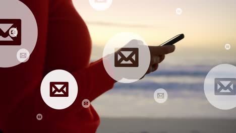 Animation-of-mail-icons-over-midsection-of-biracial-woman-using-smartphone-at-beach