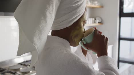 Fröhliche-Afroamerikanische-Frau-Mit-Schönheitsmaske-Im-Gesicht,-Die-In-Der-Küche-Kaffee-Trinkt