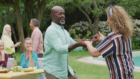 Happy-diverse-senior-female-and-male-friends-dancing-in-garden-while-having-lunch-with-friends