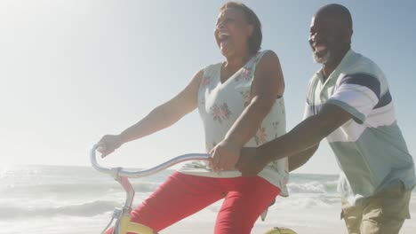 Smiling-senior-african-american-couple-riding-bicycles-on-sunny-beach