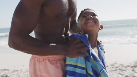 Lächelnder-Afroamerikanischer-Vater,-Der-Seinen-Sohn-Am-Sonnigen-Strand-Abtrocknet
