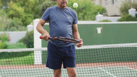 Video-of-happy-biracial-senior-man-holding-racket-and-tossing-ball-on-tennis-court