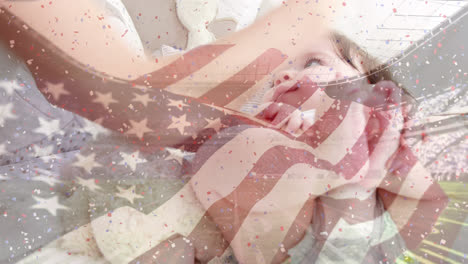 Animation-of-flag-of-usa-waving-over-stadium-and-hands-of-caucasian-mother-playing-with-baby