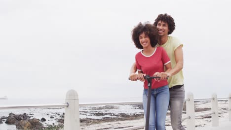 Afroamerikanisches-Paar-Lächelt,-Während-Es-Gemeinsam-Einen-Roller-Auf-Der-Promenade-In-Strandnähe-Fährt