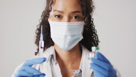 Video-Des-Porträts-Einer-Birazialen-Ärztin-Mit-Gesichtsmaske,-Die-Eine-Impfflasche-Auf-Weißem-Hintergrund-Hält