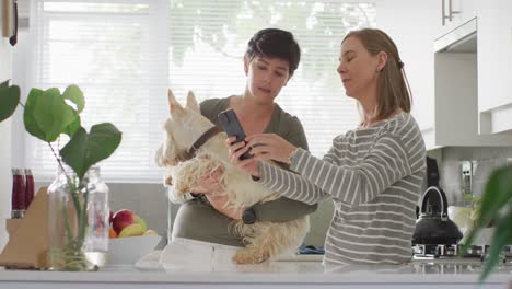 Caucasian-lesbian-couple-holding-their-dog-using-smartphone-at-home