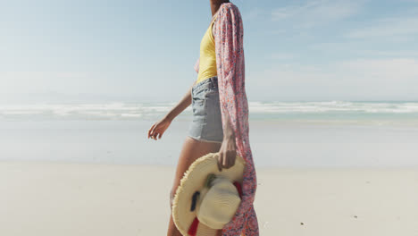 Glückliche-Afroamerikanische-Frau,-Die-Mit-Hut-Am-Sonnigen-Strand-Spaziert