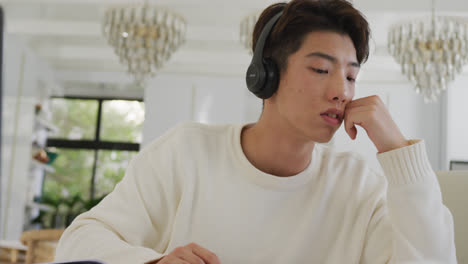 Asian-male-teenager-with-headphones-learning-and-using-laptop-in-living-room