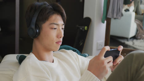 Asiatischer-Junge-Mit-Kopfhörern,-Der-Ein-Spiel-Auf-Dem-Smartphone-Spielt-Und-Zu-Hause-Auf-Der-Couch-Sitzt