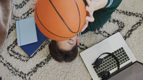 Overhead-view-of-asian-boy-playing-with-basketball-lying-on-the-floor-at-home