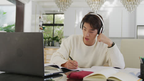 Asiatischer-Männlicher-Teenager-Mit-Kopfhörern,-Der-Im-Wohnzimmer-Lernt-Und-Einen-Laptop-Benutzt