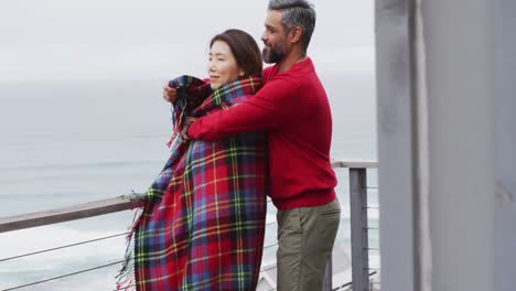 Happy-diverse-couple-with-blanket-embracing-and-talking-together-on-balcony