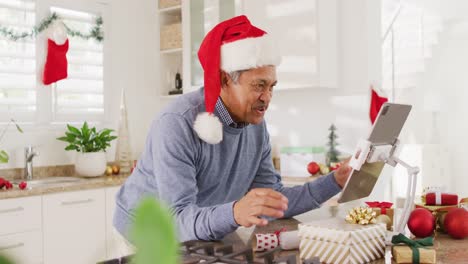 Video-of-happy-senior-biracial-man-in-santa-hat-making-christmas-video-call-on-tablet-blowing-kisses