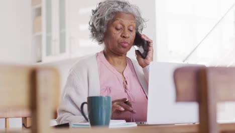 Portrait-of-senior-african-american-woman-sitting-at-table,-using-laptop-and-talking-on-smartphone