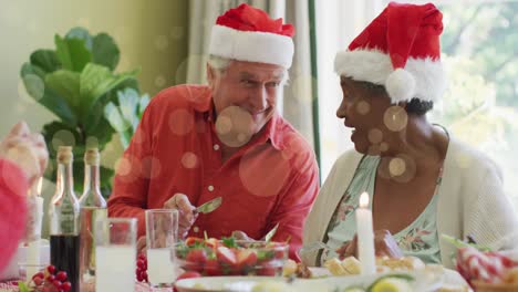 Animation-of-light-spots-over-diverse-senior-couple-in-hats-laughing-at-christmas-dinner-table