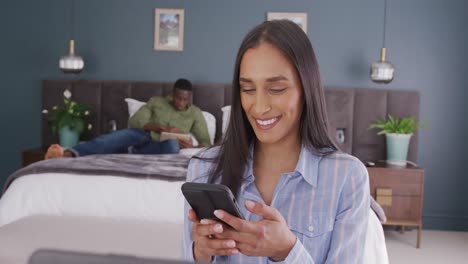 Video-of-diverse-couple-in-bedroom,-the-woman-smiling-using-smartphone-sitting-in-foreground