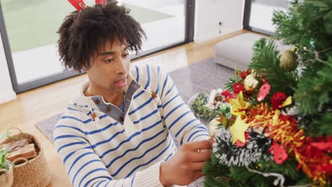 Video-of-happy-biracial-man-in-reindeer-antlers-decorating-christmas-tree-at-home