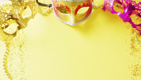 Video-of-three-masquerade-masks,-mardi-gras-beads-and-confetti-on-yellow-background-with-copy-space