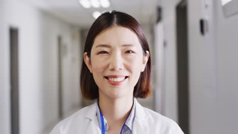 Video-portrait-of-smiling-asian-female-doctor-standing-in-hospital-corridor