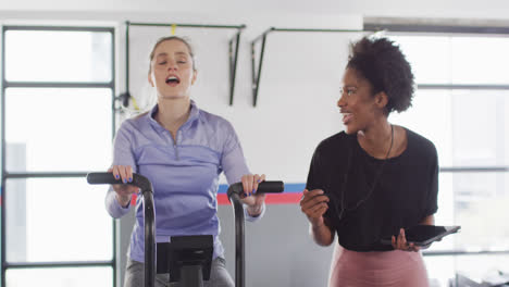 Video-of-diverse-female-fitness-trainer-and-woman-on-exercise-bike-bumping-fists-at-a-gym