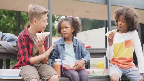 Video-of-three-diverse-schoolchildren-eating-packed-lunches,-talking-in-schoolyard