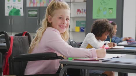 Video-portrait-of-smiling-caucasian-schoolgirl-in-wheelchair-at-desk-in-diverse-class