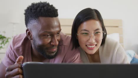Happy-diverse-couple-using-laptop-and-lying-in-bedroom