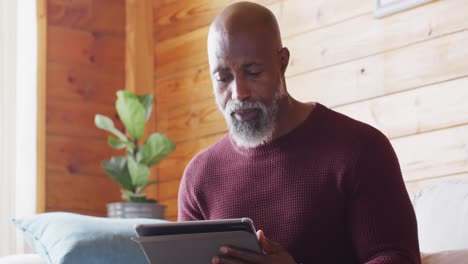 Happy-senior-african-american-man-spending-time-in-log-cabin,-sitting-on-sofa-and-using-tablet