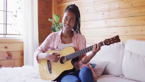 Feliz-Mujer-Afroamericana-Sentada-En-El-Sofá-Y-Tocando-La-Guitarra-En-Una-Cabaña-De-Madera,-Cámara-Lenta