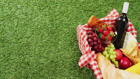 Video-of-fresh-bread,-grapes-and-wine-in-basket-and-gingham-tablecloth-with-copy-space-on-grass