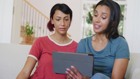 Happy-biracial-sisters-sitting-on-sofa-and-using-tablet,-in-slow-motion