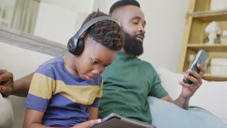 Happy-african-american-father-and-son-sitting-on-sofa,-using-smartphone-and-tablet,-in-slow-motion