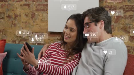 Animation-of-social-media-icons-over-diverse-couple-taking-a-selfie-from-a-smartphone