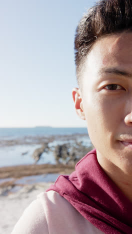 Vertical-video-of-portrait-of-happy-asian-man-looking-at-camera-at-promenade-by-the-sea,-slow-motion
