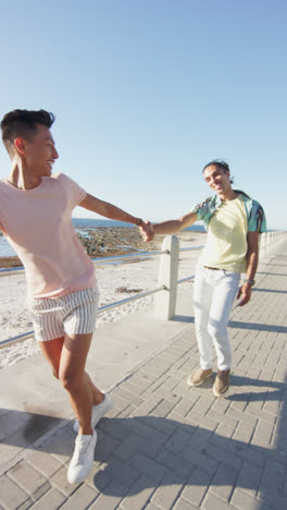 Vertical-video-of-happy-diverse-male-couple-holding-hands-at-promenade-by-the-sea,-slow-motion