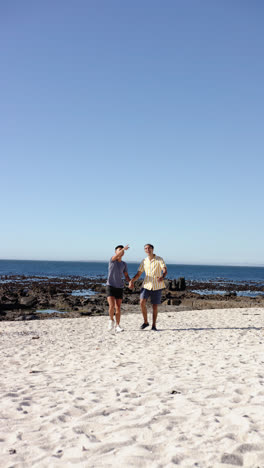 Vertical-video-of-diverse-male-couple-walking-and-holding-hands-at-beach,-slow-motion