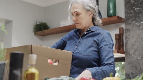 Glückliche-ältere-Kaukasische-Frau,-Die-In-Der-Küche-Eine-Kiste-Mit-Gemüse-Und-Obst-Auspackt,-Zeitlupe