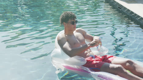 Portrait-of-relaxed-biracial-man-with-sunglasses-lying-on-inflatable-in-swimming-pool-on-sunny-day