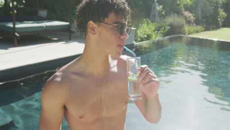 Happy-biracial-man-drinking-drink-at-pool-in-garden-on-sunny-day