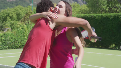 Happy-biracial-couple-with-tennis-rackets-embracing-in-garden-on-sunny-day