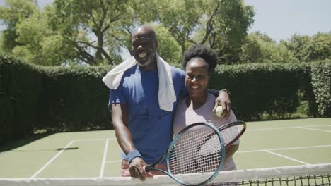 Portrait-of-happy-senior-african-american-couple-with-tennis-rackets-and-ball-at-tennis-court
