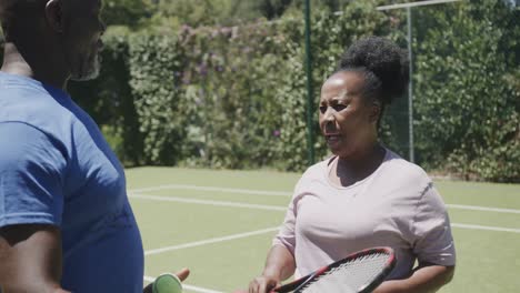 Happy-senior-african-american-couple-with-tennis-rackets-talking-at-tennis-court-in-slow-motion