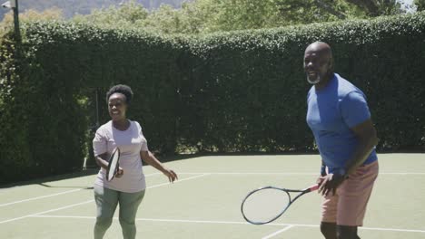 Happy-senior-african-american-couple-playing-tennis-and-high-fiving-at-tennis-court-in-slow-motion