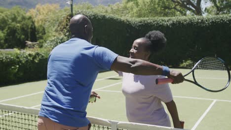 Happy-senior-african-american-couple-playing-tennis-and-embracing-at-tennis-court-in-slow-motion