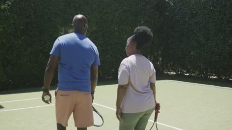 Happy-senior-african-american-couple-walking-with-tennis-rackets-at-tennis-court-in-slow-motion