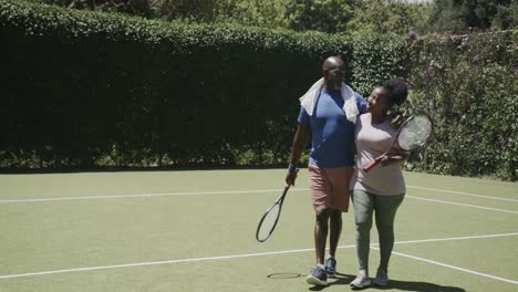 Happy-senior-african-american-couple-walking-with-tennis-rackets-and-embracing-in-slow-motion