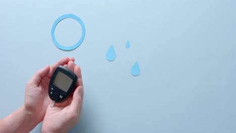 Hands-of-caucasian-woman-holding-glucometer-over-blue-drops-and-circle-on-pale-blue,-slow-motion